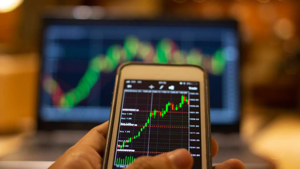 Hand holding a smartphone displaying a stock market chart, with a laptop showing a similar chart blurred in the background.