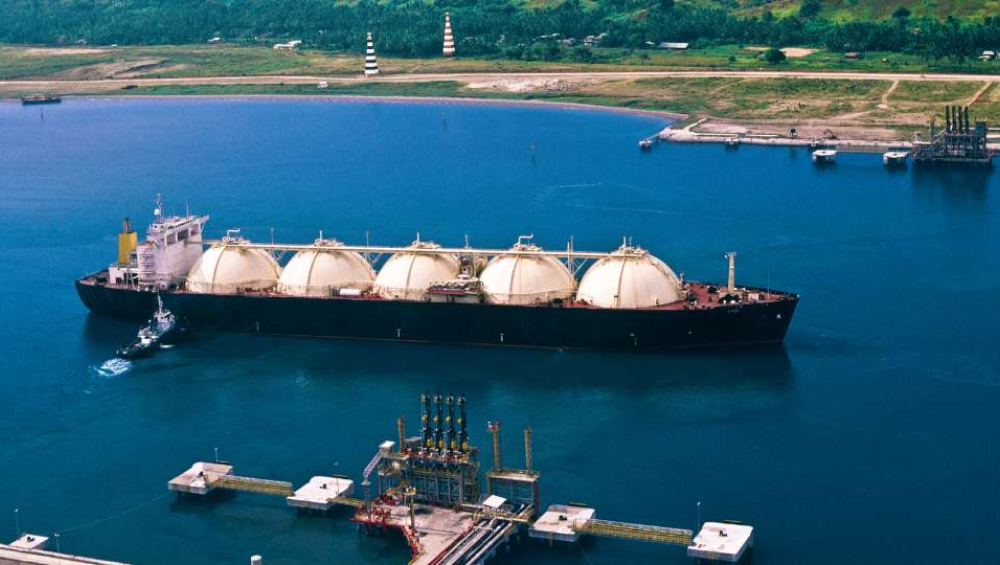 Large LNG carrier ship with four spherical tanks being assisted by a tugboat at an industrial terminal.