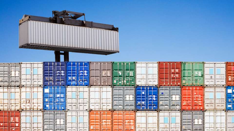 Industrial port crane lifting a grey shipping container onto a stacked wall of containers.
