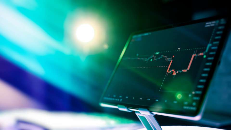 Tablet displaying a red and green candlestick chart on a dark background, set against dramatic blue lighting.