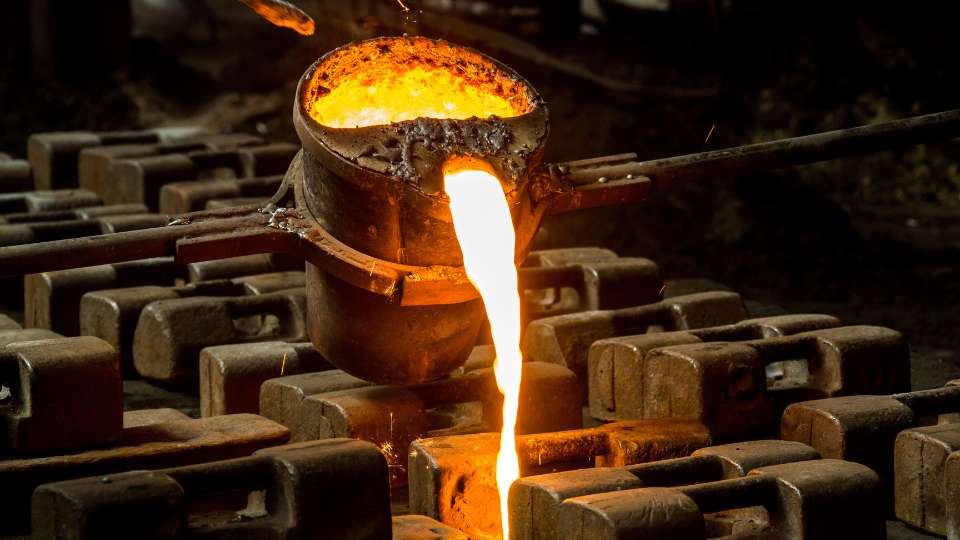 Glowing orange molten metal pouring from a ladle into molds in a foundry.