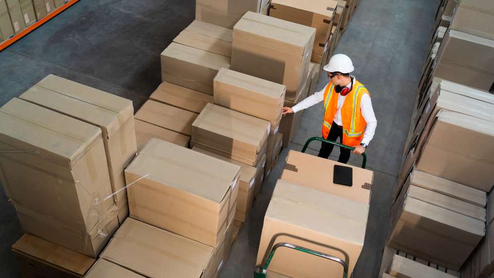 Warehouse worker in a hard hat moving boxes with a hand truck.