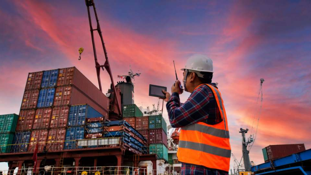 Logistics supervisor using a radio and tablet in front of shipping containers at sunset.