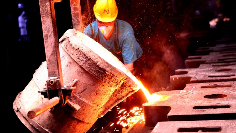 Industrial worker in protective gear pouring molten metal from a ladle in a foundry.