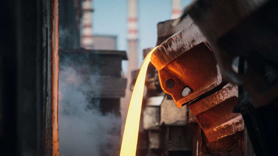 Molten steel pouring from a ladle in a steel mill with industrial smokestacks visible.