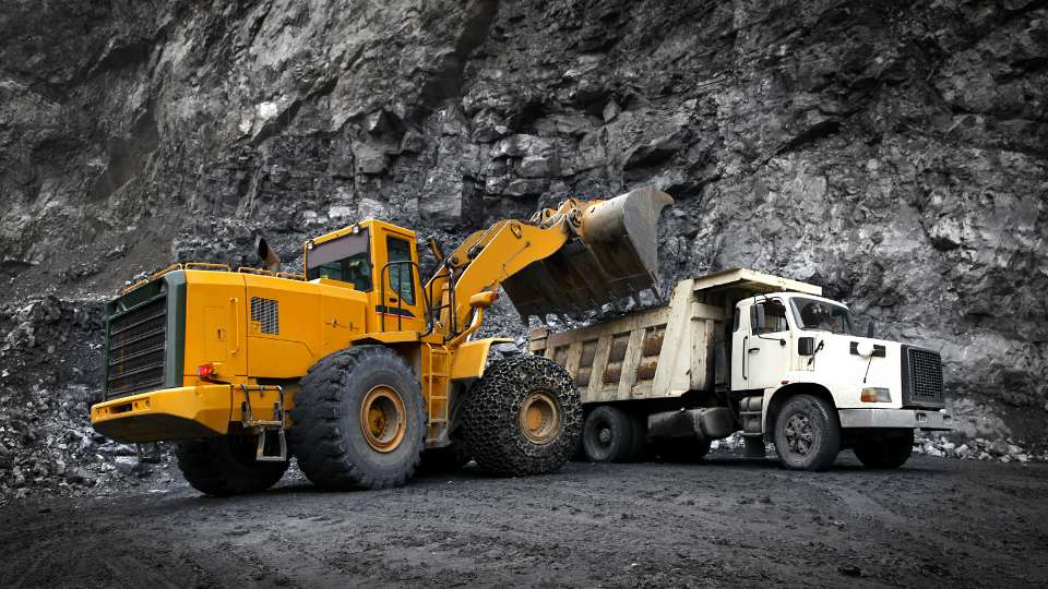 Large yellow front-end loader filling a heavy-duty dump truck with dark coal or mining aggregate against a rocky backdrop.