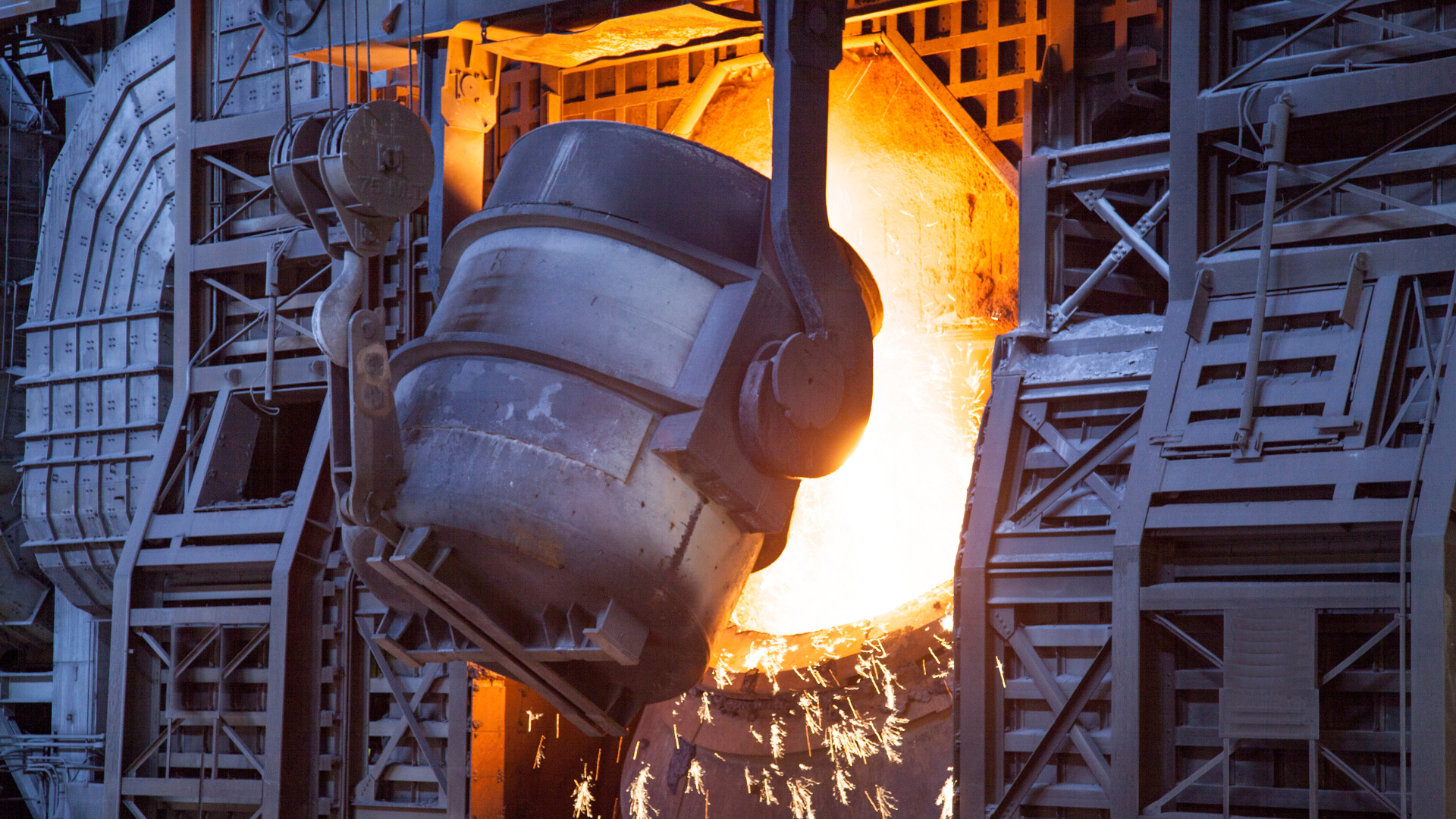 A large ladle pouring molten metal in a dramatic, sparks-flying steel mill environment.
