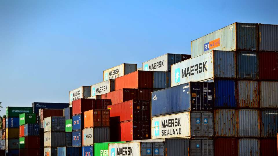 Stacked multicolored shipping containers marked with major logistics company names like MAERSK and Evergreen, in a busy port or storage yard.