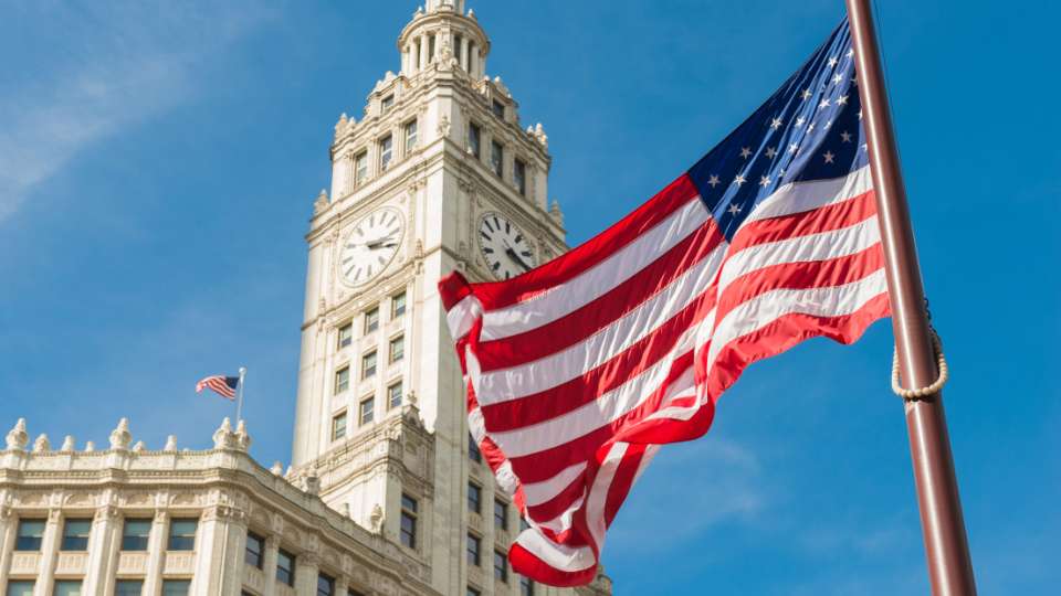 Large American flag waving in the wind in front of the historic Wrigley Building clock tower in Chicago.