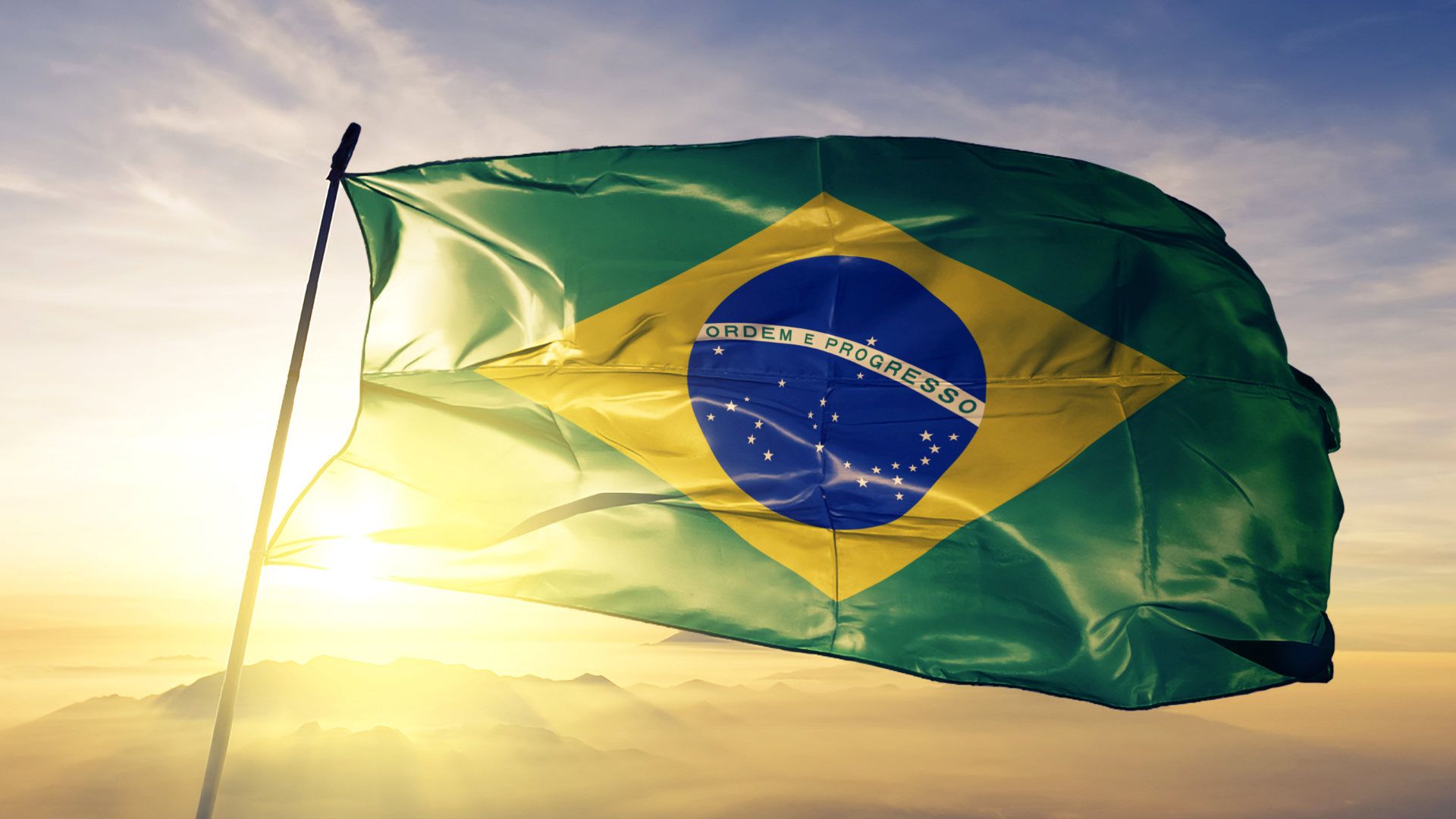 Brazilian national flag waving against a dramatic sunset and cloudy sky over mountains.