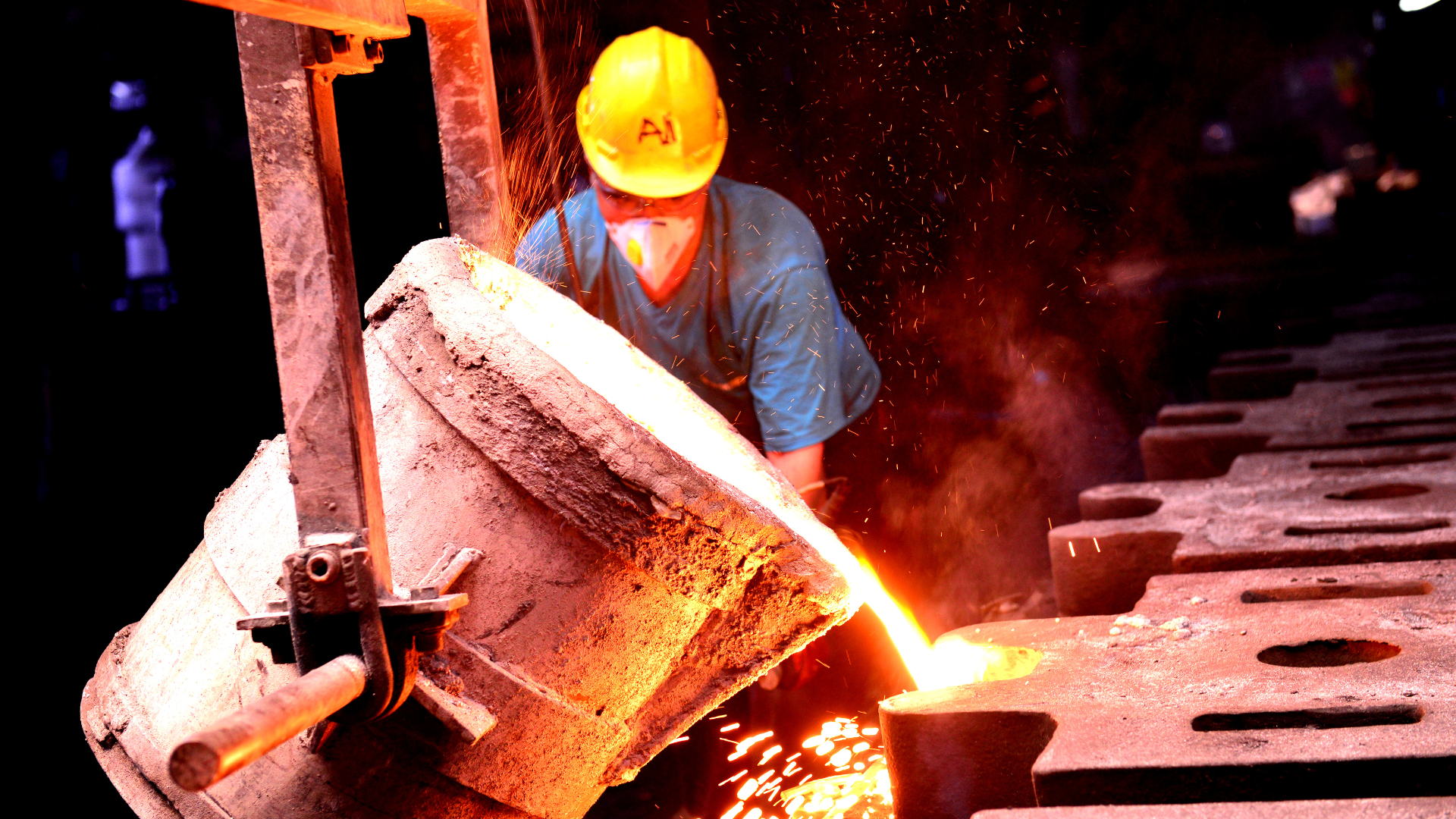 Industrial worker wearing a yellow hard hat pouring molten metal from a crucible in a foundry.
