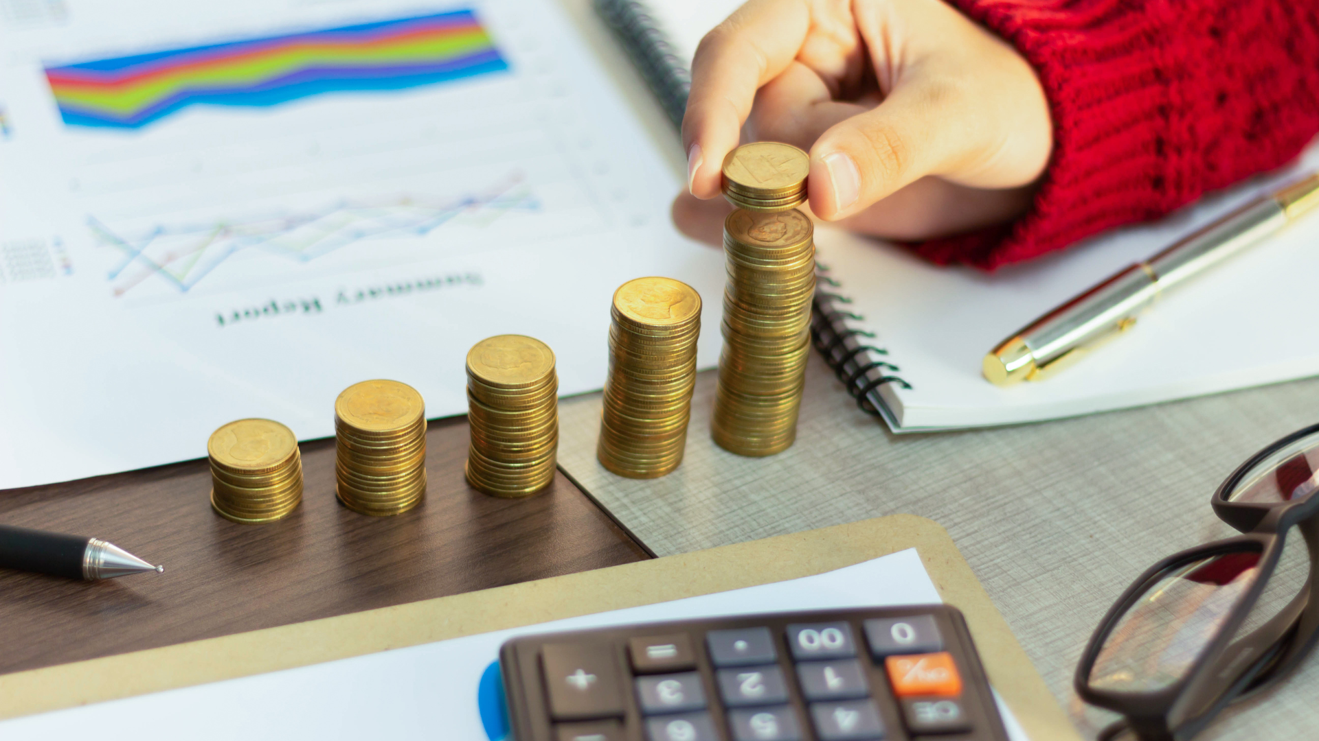 Hand stacking gold coins into bar graphs next to financial charts, symbolizing growth.