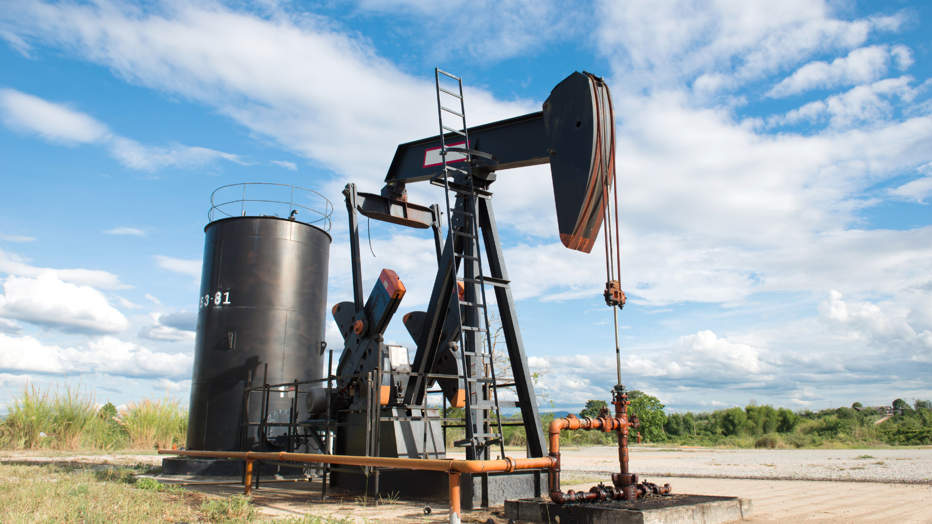 Oil pump jack extracting crude oil in a sunny field with blue sky and storage tank nearby.