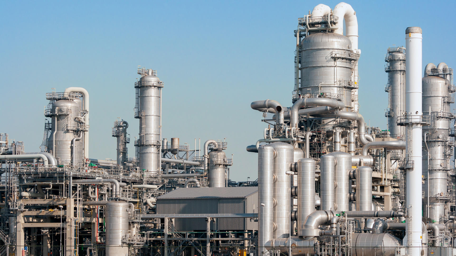 Large industrial oil and gas refinery with metal pipelines, storage tanks, and distillation towers under a clear blue sky.