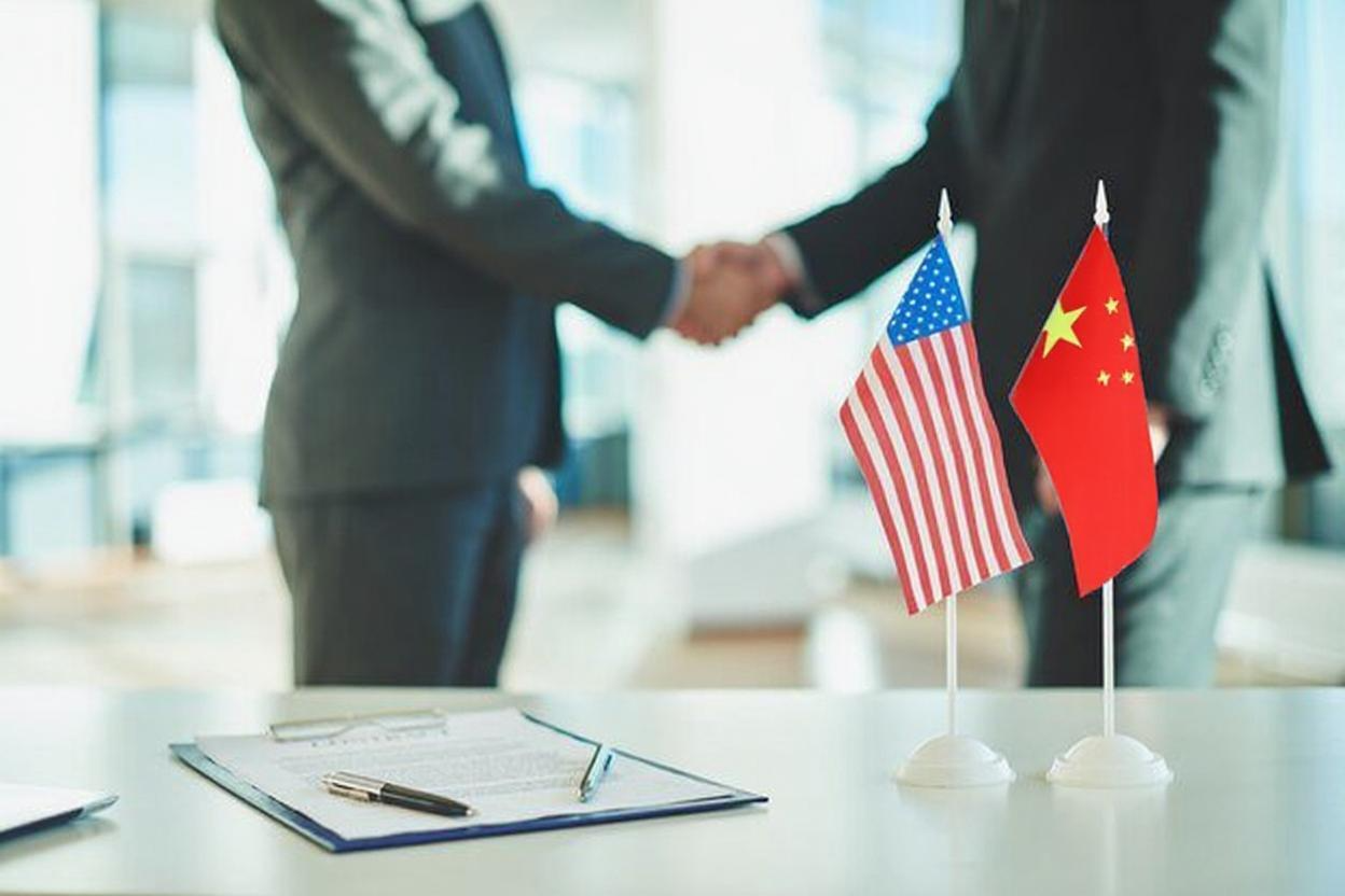 Two businessmen shaking hands over a contract, with US and China miniature flags on the table.