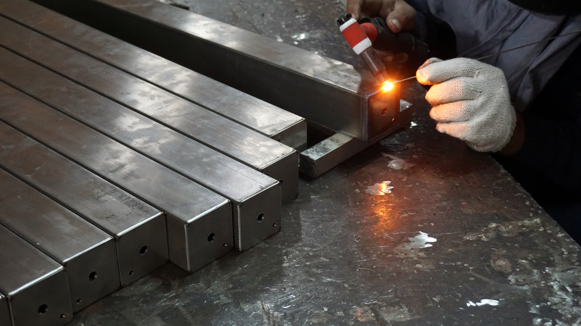 Worker performing TIG welding on stainless steel rectangular tubes in a metal workshop.
