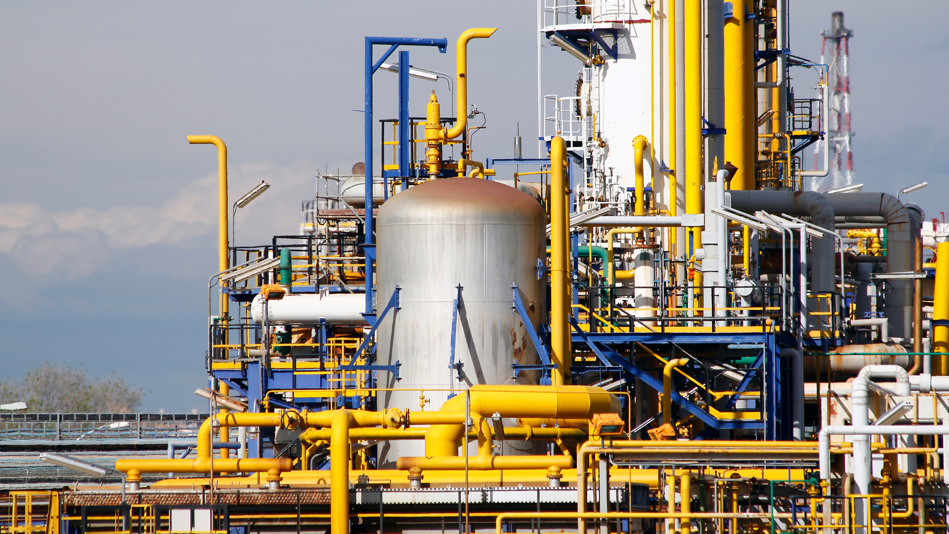 Industrial oil and gas refinery with metal tanks, yellow pipelines, and processing equipment under a clear sky.