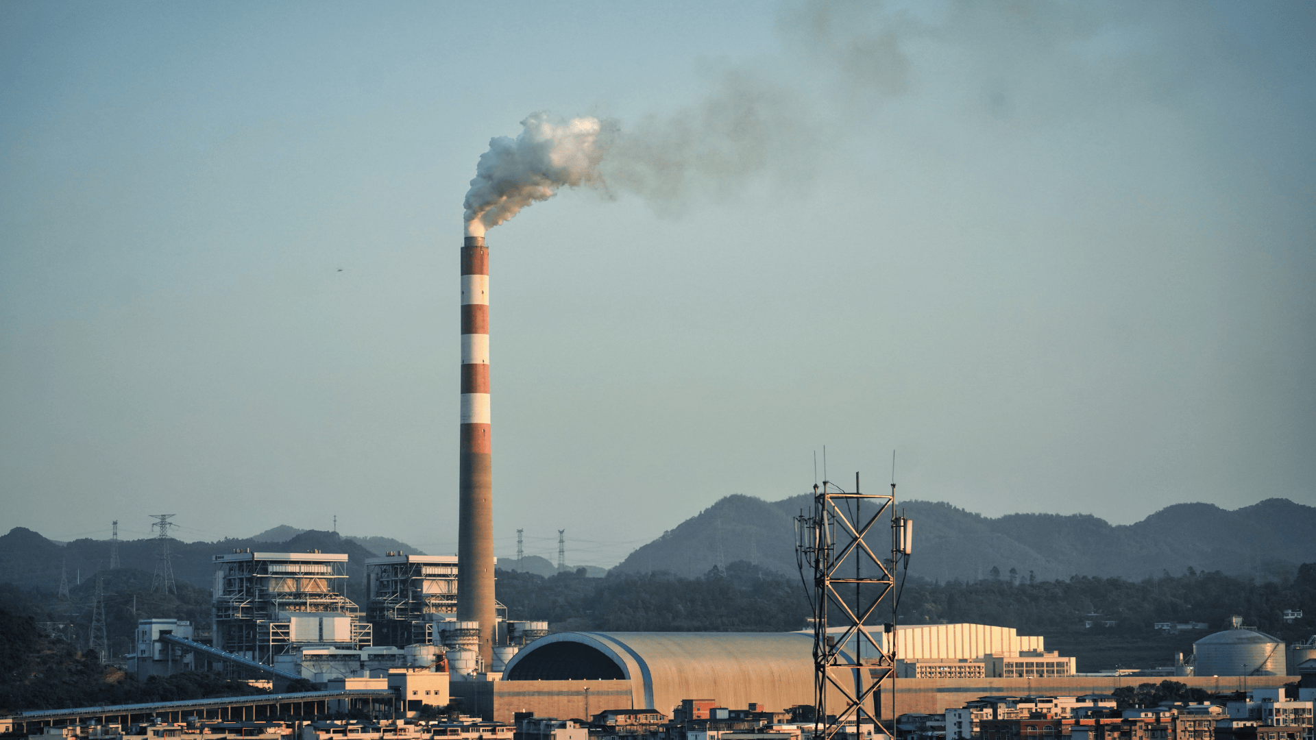 Industrial smokestack emitting smoke at a power plant with mountains in the background.