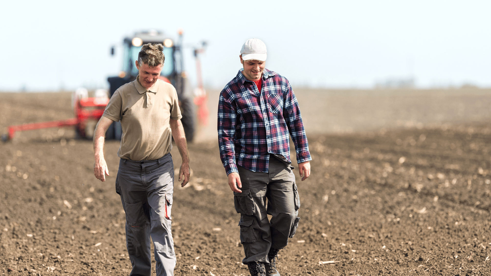 Two farmers walking through a freshly plowed field with a tractor in the background.
