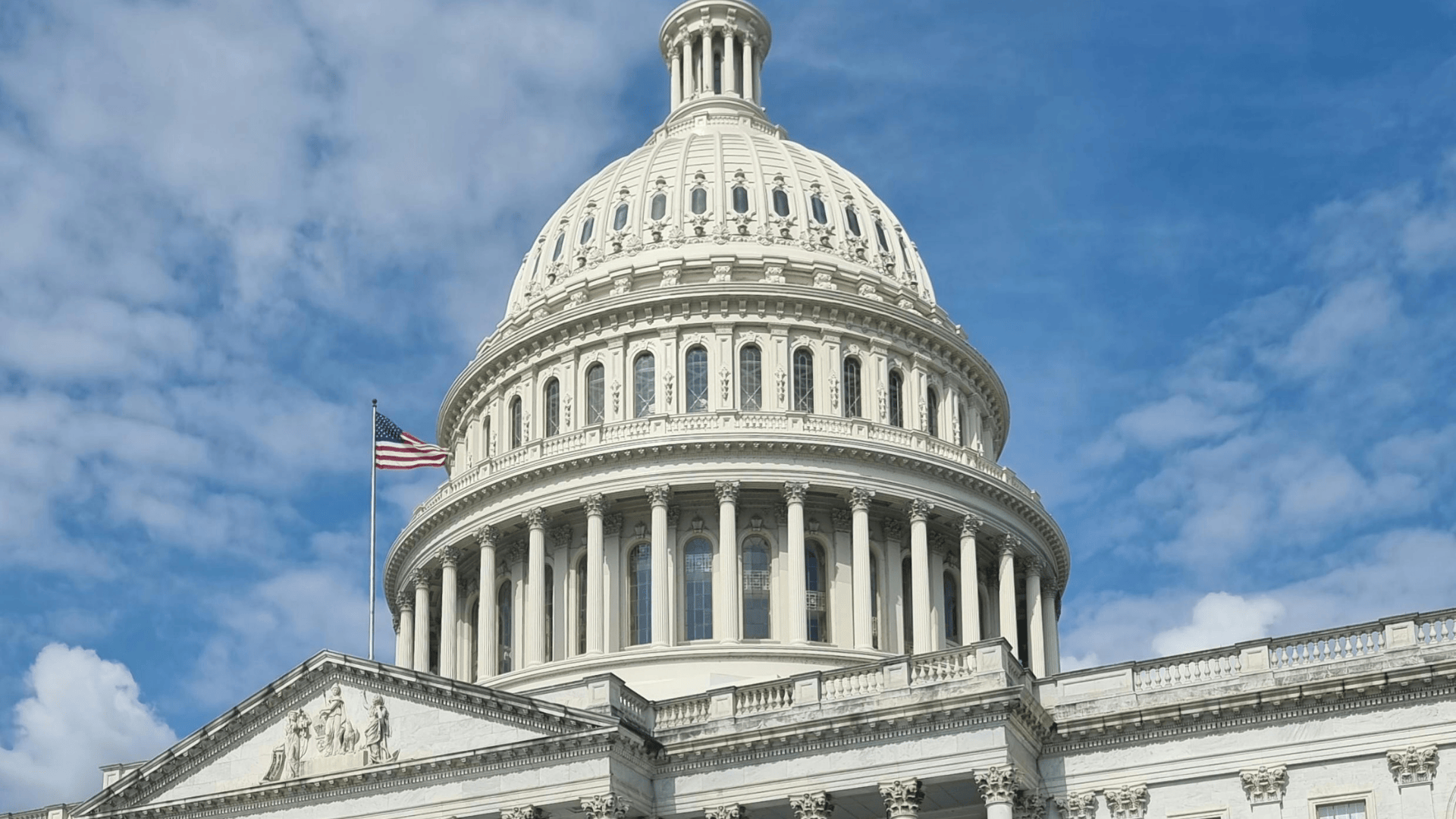 Close-up of the United States Capitol Building dome with the American flag flying against a blue, cloudy sky.