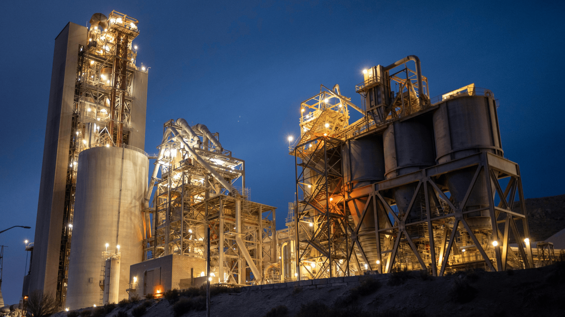 Industrial cement plant illuminated at night with large silos, towers, and metal structures under a deep blue sky.