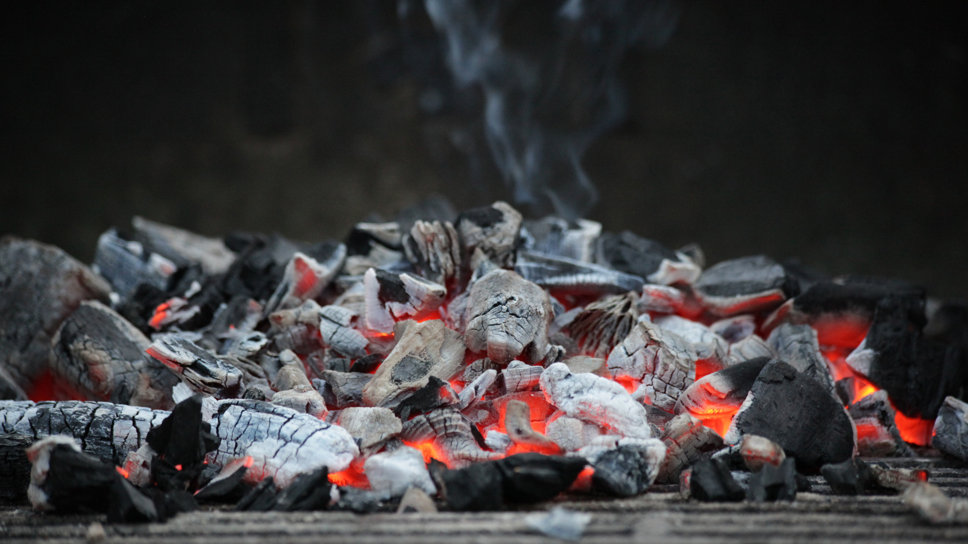 Glowing charcoal embers with ash and light smoke, showing burning coals used as an energy or fuel source.